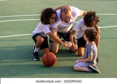 Coach And Group Of Kids Discussing Tactics On The Basketball Court.Young Trainer Giving His Students Talk On The Green Field.Basketball Coach Discusses Play With His Team With Their Hands In Huddle.