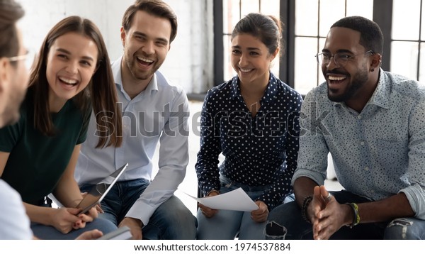 Coach or group leader telling funny story or joke\
to laughing diverse team. Happy multiethnic employees having fun\
while discussing project, sharing ideas, informal brainstorming\
sitting on chairs