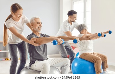 Coach or health care worker helping mature people to do sport exercises with dumbbells sitting on fit ball in gym. Physiotherapist helping retired man and woman in rehab in rehabilitation center. - Powered by Shutterstock