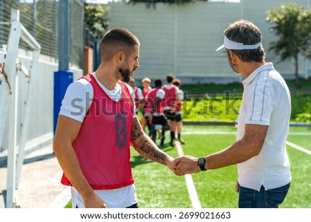 Coach and young soccer player shaking hands on a green sports field, demonstrating teamwork, respect, and sportsmanship during training with other teammates in the background