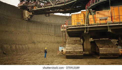 Coal Mine Worker With A Helmet On His Head Standing In Front Of Huge Drill Machine And Looking At It. Rear View.