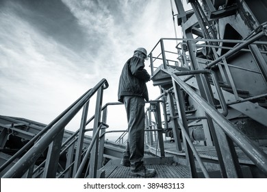 Coal Mine Worker With A Helmet On His Head Standing In Front Of Huge Drill Machine And Looking At It. Side View. Image Is Carefully Post Processed And Toned.