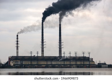 Coal Power Plant High Pipes With Black Smoke Moving Upwards Polluting Atmosphere With Reflections Of It In Lake Water.