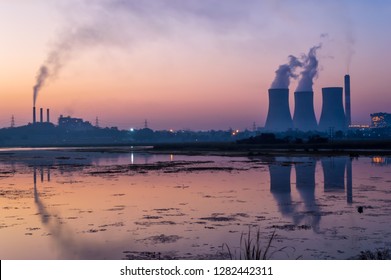 Coal powered thermal power plant. Chimney emitting smoke and cooling tower emitting steam. The lake near the electricity generating power plant showing reflections.