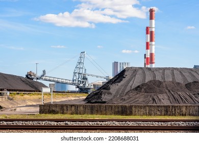 Coal Stockpiles And Bucket-wheel Stacker-reclaimer In A Thermal Power Plant With Red And White Smokestacks On A Sunny Day.