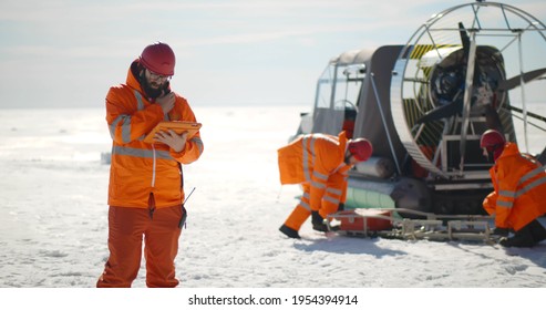 Coast Guard Team Standing On Frozen Lake With Hovercraft On Background. Rescuers In Safety Uniform Loading Equipment In Airboard Preparing To Patrol Arctic Area