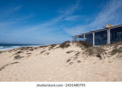 A coastal beachscape featuring a modern structure with large glass windows nestled in the sand dunes of Comporta, Setubal, Portugal, under a blue sky - Powered by Shutterstock