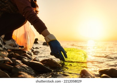 Coastal Cleanup And Garbage Collection For Recycling. A Woman Volunteer Collects Plastic Bottle By The Sea Or River, Closeup Of Hand. Low Angle View. World Environment Day And Earth Day Concept.