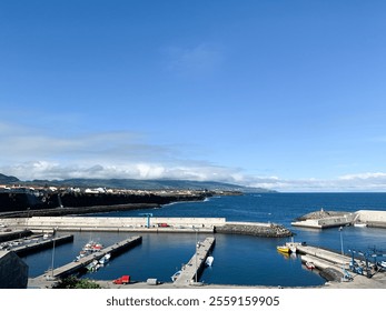 Coastal harbor with docks, fishing boats, and blue ocean under clear sky. Green hills and village buildings in the background. Captured in Rabo de Peixe, Azores island São Miguel, Portugal. - Powered by Shutterstock