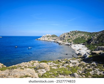 A coastal landscape with a clear blue sky, calm sea, rocky shoreline, and boats floating near the shore - Powered by Shutterstock