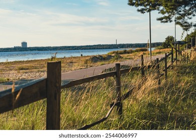 A coastal path bordered by a wooden fence runs alongside a grassy shoreline, offering a peaceful and scenic view of the ocean under a bright blue sky - Powered by Shutterstock