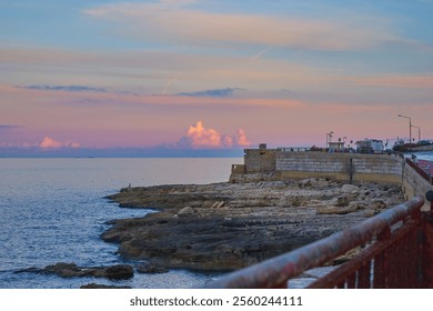 Coastal promenade featuring rocky shoreline under pastel-colored sunset sky, with calm sea waters stretching into horizon - Powered by Shutterstock