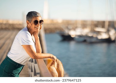 Coastal Relax. Delighted senior woman wearing her sunglasses and holding beach hat at the marina, looking at sailboats. Tourist embracing sea breeze and holiday spirit by boats, free space - Powered by Shutterstock