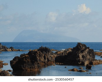 Coastal scene in the Azores, featuring rugged dark volcanic rocks against a calm blue sea, with a distant island silhouette, Corvo island, under a lightly clouded sky. - Powered by Shutterstock