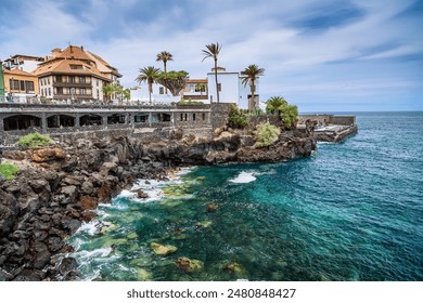 A coastal scene from Puerto de la Cruz in Tenerife featuring a rocky shoreline with clear, turquoise water. A traditional building with a terracotta roof surrounded by palm trees - Powered by Shutterstock