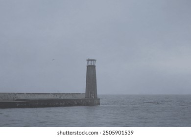 Coastal scene with a tall, narrow lighthouse on a pier extending into the sea under overcast skies. Calm waters and misty horizon create a serene maritime landscape autumn grey fall climate atmosphere - Powered by Shutterstock