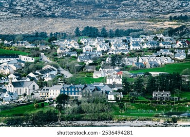 A coastal town with a diverse array of houses, a church, and a small harbor in the foreground. The town is surrounded by lush greenery and a rocky hill in the background. - Powered by Shutterstock