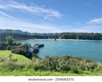 Coastal town overlooking a vibrant blue bay with colorful houses along the cliffside and a small pier extending into the water. Lush greenery in the foreground and a clear summer sky above. - Powered by Shutterstock