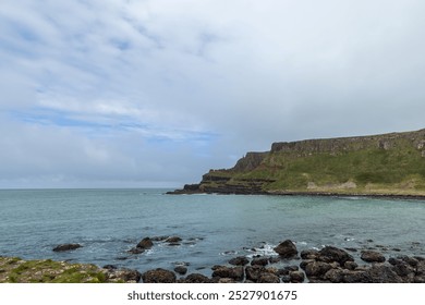 Coastal view of Giant Causeway, Northern Ireland, showing rugged basalt rocks along the shoreline, with green cliffs and the calm Atlantic Ocean under a cloudy sky - Powered by Shutterstock