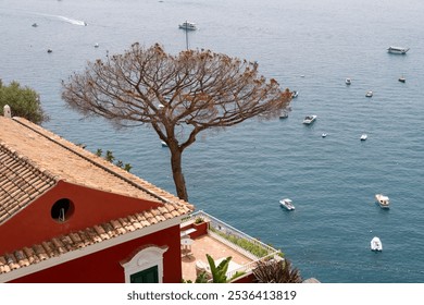 Coastal view of a Mediterranean villa with a red facade and terracotta roof, overlooking calm blue waters dotted with boats and a unique tree in the foreground - Powered by Shutterstock