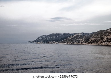 Coastal view showcasing rocky cliffs and calm waters on a cloudy day near a serene shoreline with gentle waves - Powered by Shutterstock