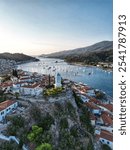 coastal village with red-tiled rooftops, centered around a charming blue-domed church tower, overlooks a serene bay filled with boats. The scenic backdrop of mountains and calm waters creates a tranqu