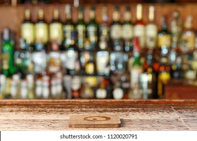 Coaster Under Glass On Blurred Bar Background. Image Of Wooden Bar Counter In Front Of Abstract Blurred Background Of Alcohol In Resturant 