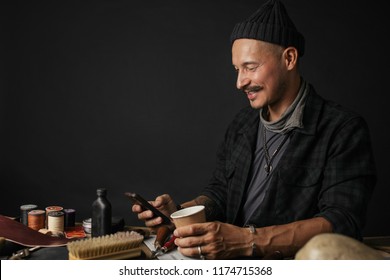 Cobbler Man using smart phone and holding cup of coffee in front of table with tools at shoemaking workshop - Powered by Shutterstock