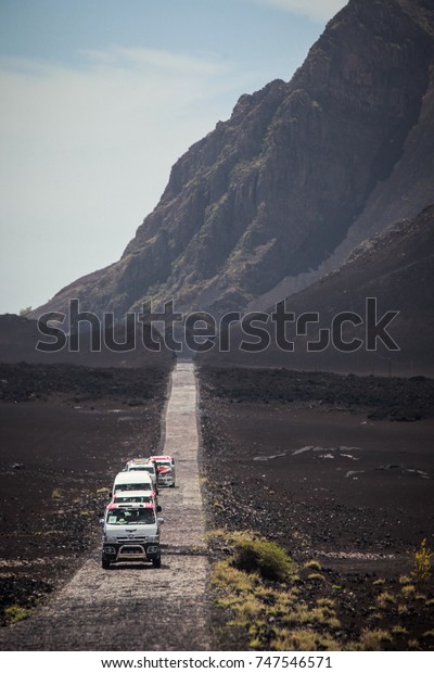 Cobblestone road\
with a caravan of cars and vans on it. Hills in the background.\
Island of Fogo, Cabo\
verde.