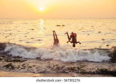 Cochin, India - November 22, 2015: Too Crazy Friends Jumping Through Big Wave In Sea. Indian People Having Rest On The Local Beach At Sunset In Fort Kochi, Kerala State