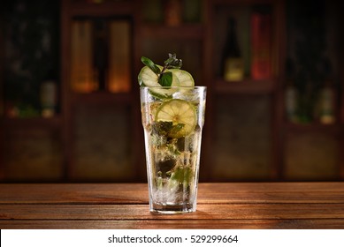 Cocktail with alcohol, lime slices, herbs and ice cubes on a wooden table with blurred background of bottles and light. 