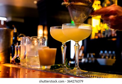 Cocktail Being Poured At A Bar. Motion Blur Of The Bartender's Hand And Strainer. Selective Focus On The Foreground Glass.