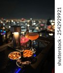 A cocktail and a bowl of nuts placed on a table at a rooftop bar of a high skyscraper. The vibrant city lights twinkle in the background, creating a stunning night-time atmosphere.