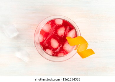 A Cocktail With Campari And An Orange Curl Garnish, Shot From Above On A White Wooden Background With Ice Cubes And A Place For Text