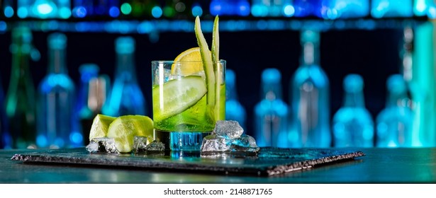 Cocktail with cucumber, ice on bar counter in a restaurant, pub. Fresh tonic drink with lime juice, mint, gin, cucumber juice. Alcoholic cooler beverage at nightclub on dark background.