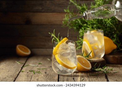 Cocktail gin tonic with ice, lemon, and rosemary on a old wooden table. Gin is poured from a bottle into a glass with ice and lemon slices. Copy space.
