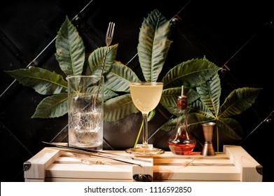 Cocktail Glass With Yellow Summer Cocktail, Bottle With Red Liquor And Bar Utensils Arranged On The Background Of Green Leaves And Black Wall On Wooden Box