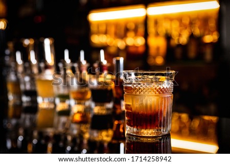 A cocktail in an old fashioned glass on a bar counter with a reflection, bottles with bitters, bokeh lights, horizontal photo