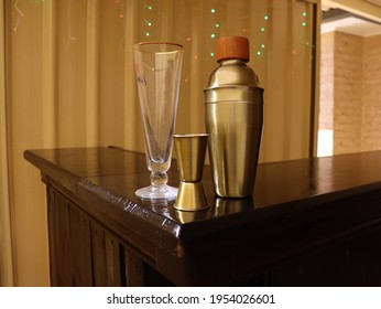 Cocktail Shaker, Glass And Measuring Cup On Glossy Brown Varnished Home Made Bar Top In Suburban Backyard Setting.