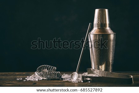 Cocktail shaker, swizzle, tongs and spoon with crushed ice for preparing a summer cocktail beverage on an old vintage wooden table with copy space