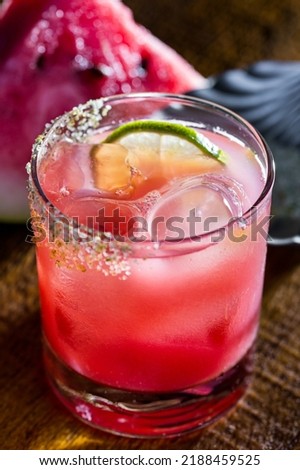 A cocktail with syrup, ice and lime in a glass that stands on the edge of a wooden table against a background of watermelon