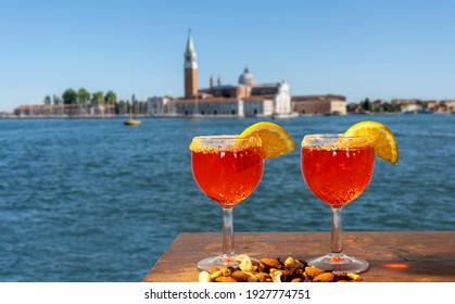 Cocktail For Two With Peanuts Snacks On Wooden Table With Venice Panorama In The Background. Spritz With Orange Slice.