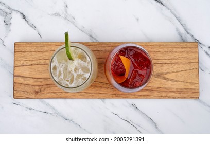 Cocktails. Top View Of A Tropical Caipirinha Drink With Ice And Lime, And A Negroni Cocktail With Orange Peel, On A Wooden Tablet On The White Marble Table. 