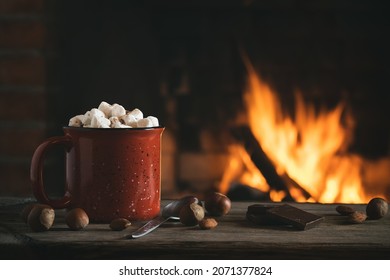 Cocoa with marshmallows and chocolate in a red mug on a wooden table near a burning fireplace - Powered by Shutterstock