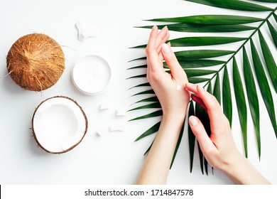 Coconut oil hand cream, organic cosmetic for skin care concept. Female hands applying coconut lotion or moisturizer over white background with coconuts and tropical palm leaf. Flat lay, top view