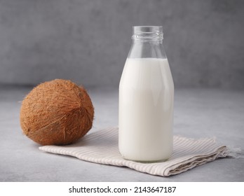 Coconut Vegan Milk Is Non Dairy Products In A Bottle With A Place To Copy On A Gray Concrete Background. Glass Bottle Of Milk Or Yogurt With Coconut Chips Aside. Concept Of Healthy Eating. Close Up.