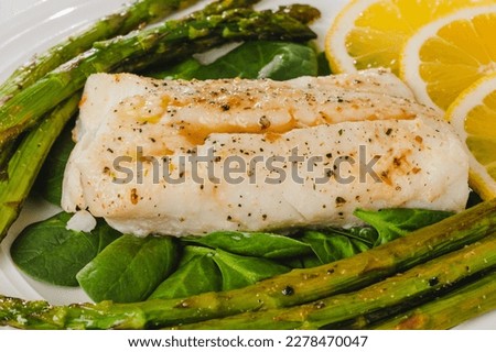 Cod fillet baked with garlic butter sauce served with fresh spinach and roasted asparagus on a white plate close-up on the wooden background, flat lay with copy space