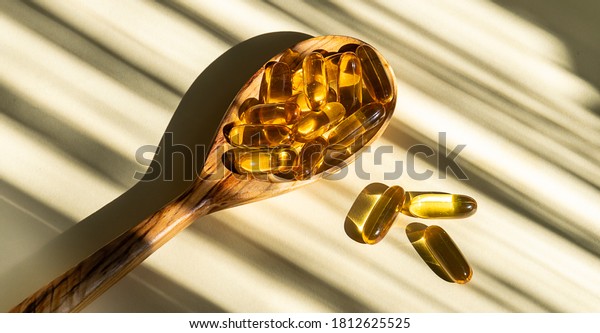 Cod liver oil capsules with vitamin D in the\
wooden spoon on light beige background with hard shadows. Health\
care concept.