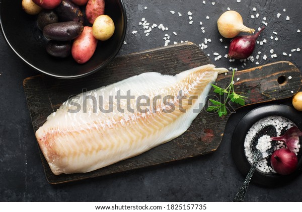 Cod white fish potatoes dish ingredients for\
healthy comfortable home food. Raw white fish fillet in a baking\
dish on a dark background. Top\
view.