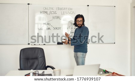 Code academy online. Young serious bearded male teacher wearing glasses pointing at whiteboard and teaching HTML CSS, giving lesson online. Focus on a man. E-learning. Distance education. Stay home